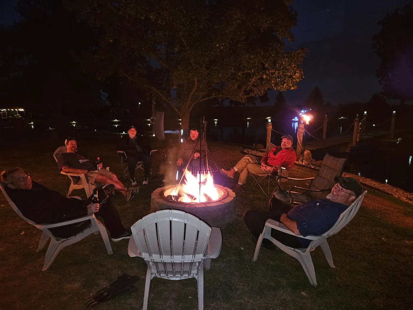 Friends relaxing in chairs around a lit fire pit at night, the pond and dock lights glowing behind them.