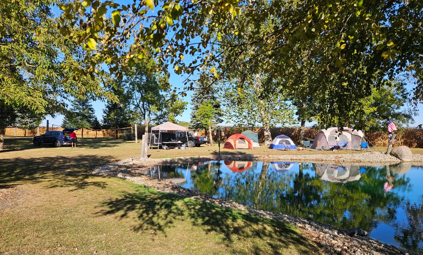 Tents pitched on the grass beside the pond under a clear blue sky, with an American flag and a cornfield beyond.